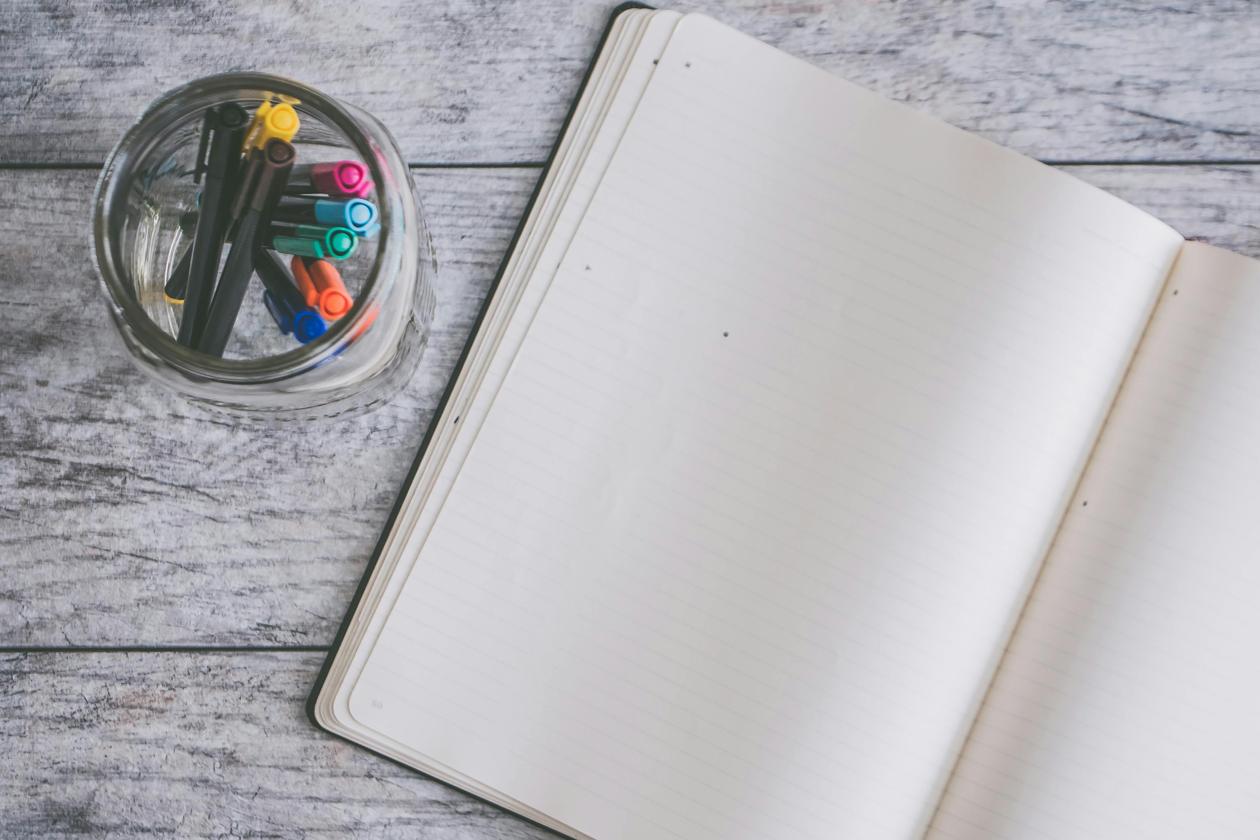 An open notebook and colorful pens in a glass jar on a wooden desk.