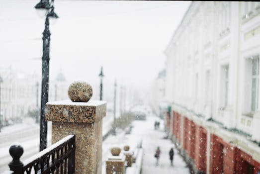 Blurred view of a snowy urban street with red brick buildings and falling snowflakes.