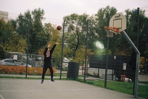 A young man is shooting a basketball on an outdoor court during autumn, surrounded by trees.
