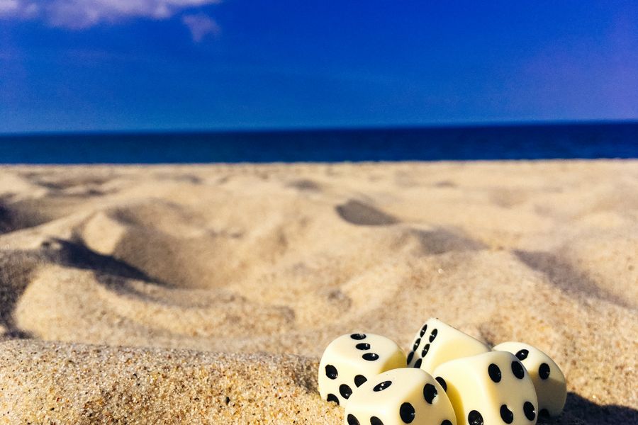Five dice resting on a sunlit beach with ocean in the background, perfect for summer and leisure themes.