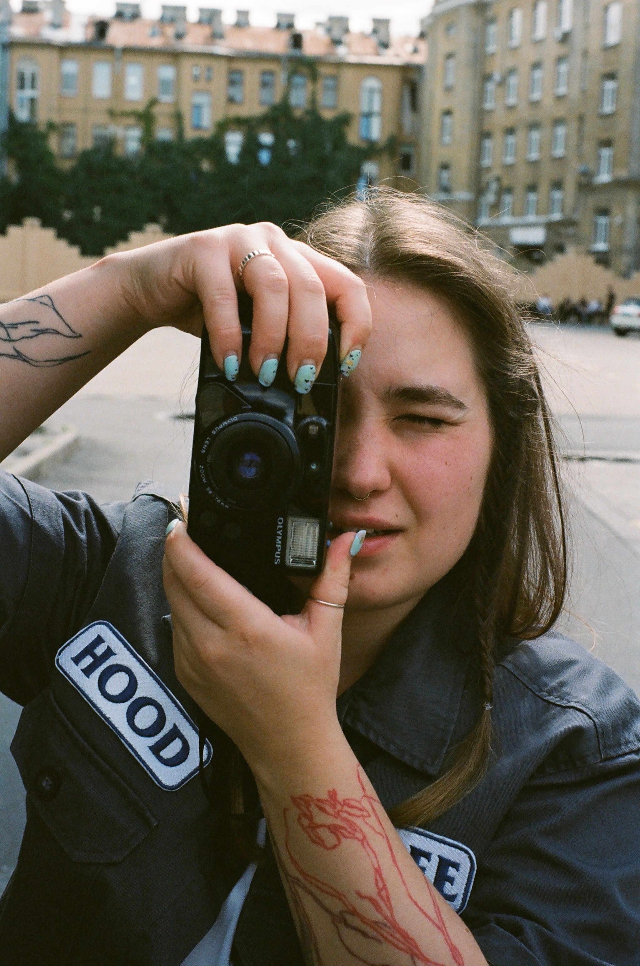 A woman with tattoos and long brown hair takes a photo in a bustling city environment.