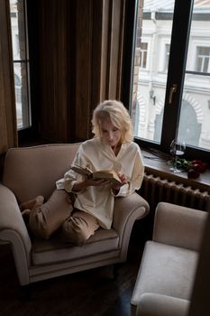 Senior woman reading a book in an armchair by the window indoors.. senior, woman, reading