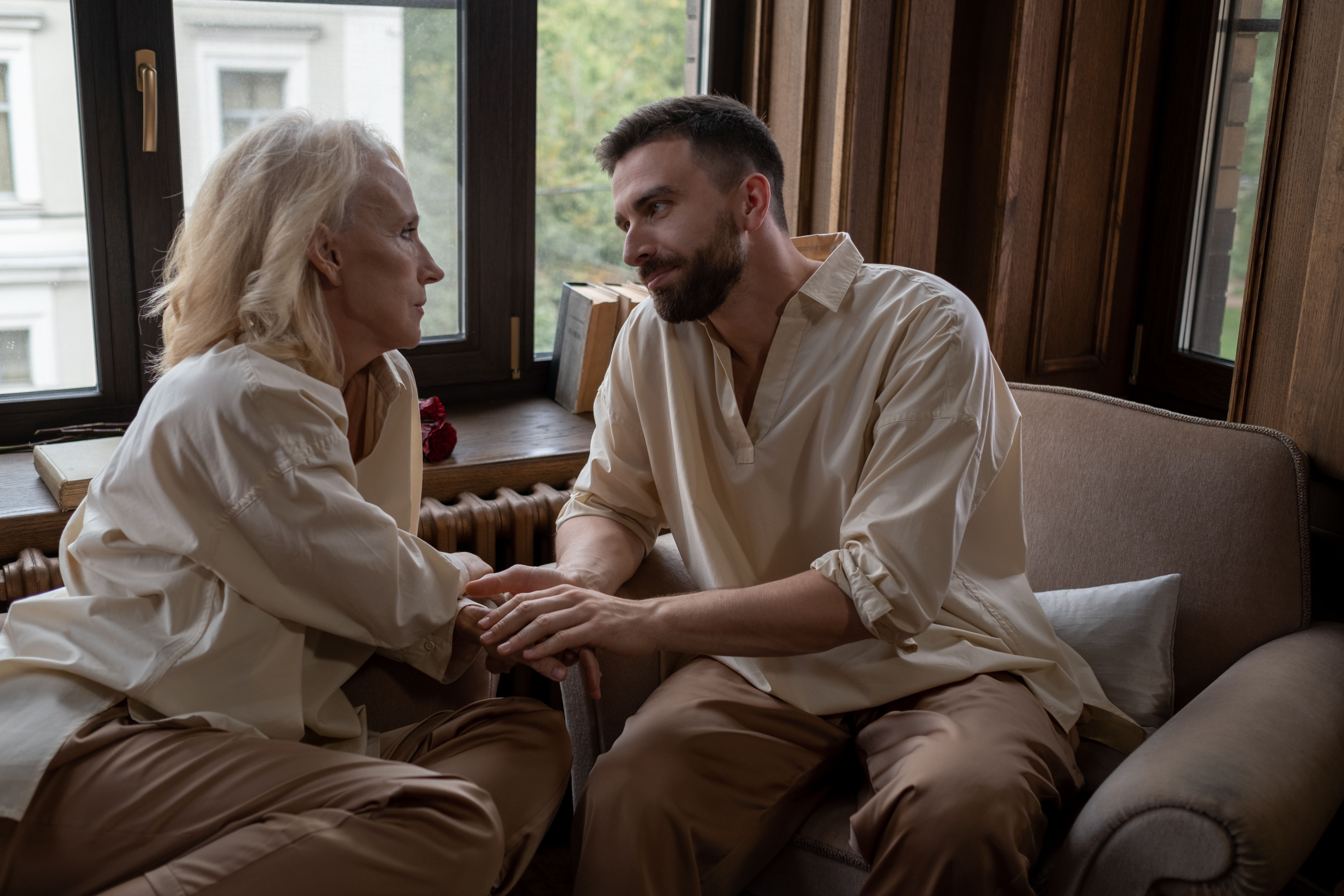 An elderly woman and a young man share a heartfelt moment by the window indoors.