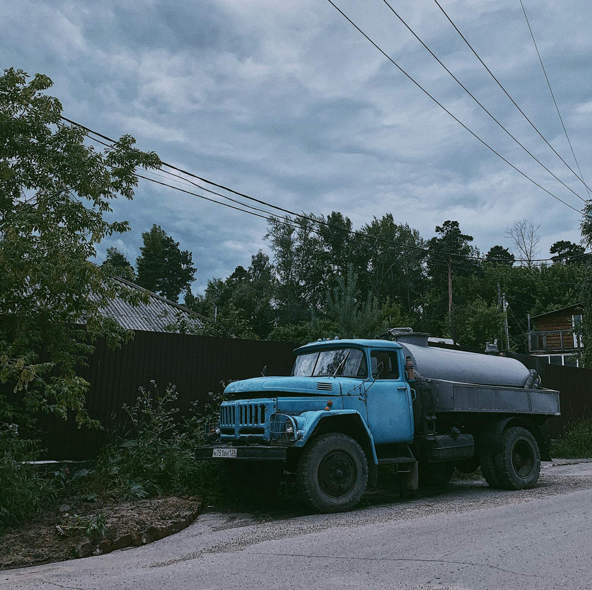 A vintage blue tanker truck parked on a rural street in Irkutsk, Russia, with lush green surroundings.