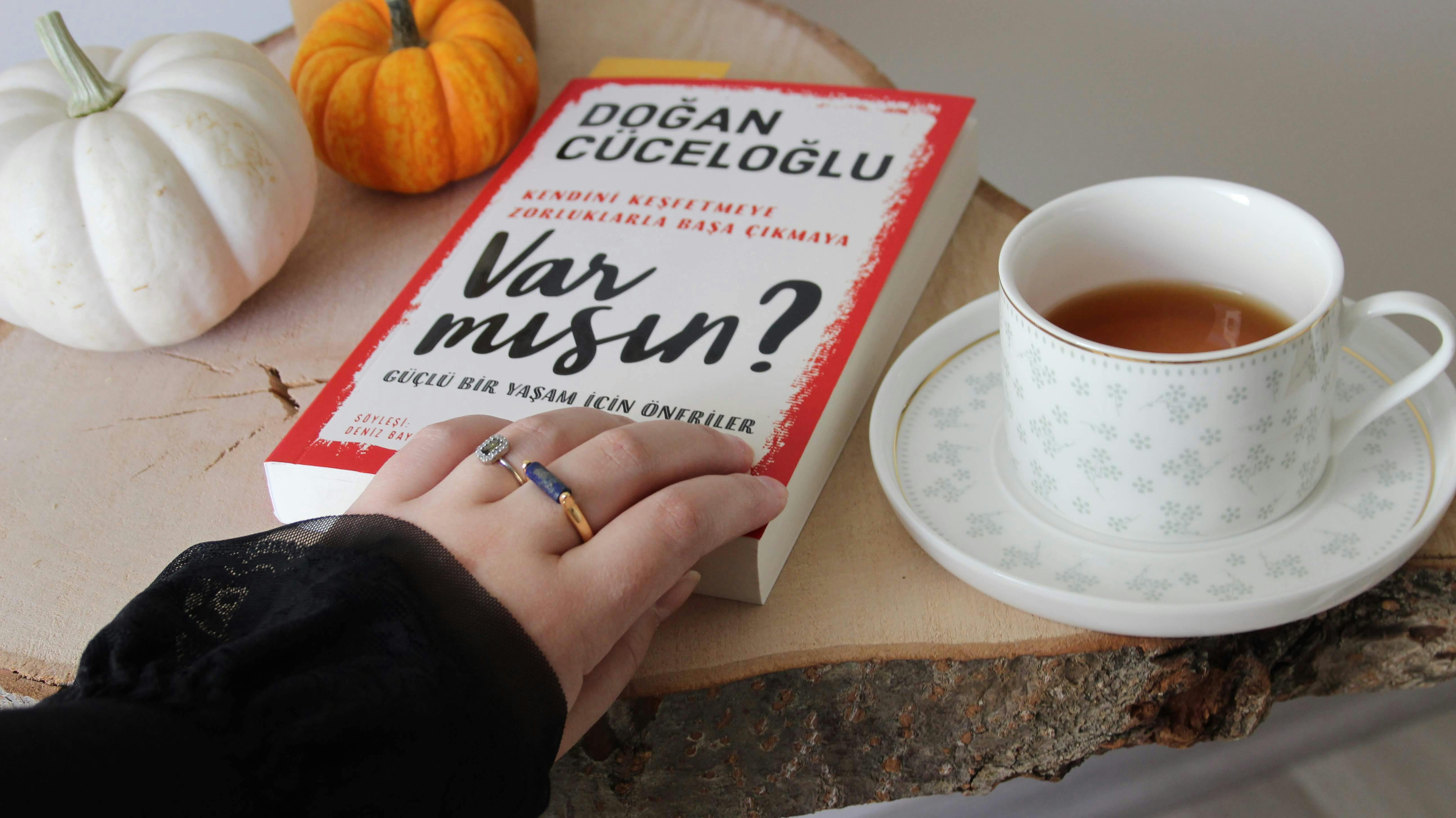 A hand with rings holds a book beside a tea and pumpkins on a wooden table.