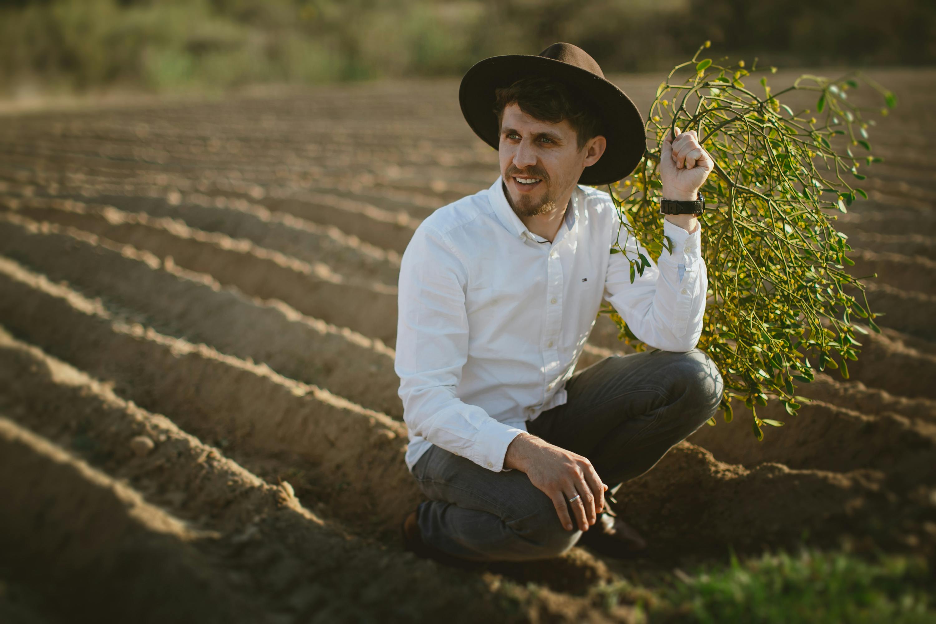 Man wearing a hat crouching in a plowed field holding mistletoe.