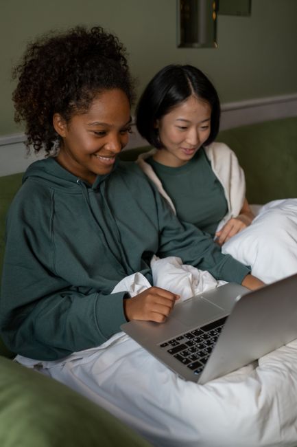 Two young women sitting on a sofa, smiling as they look at a laptop screen, enjoying their time together.