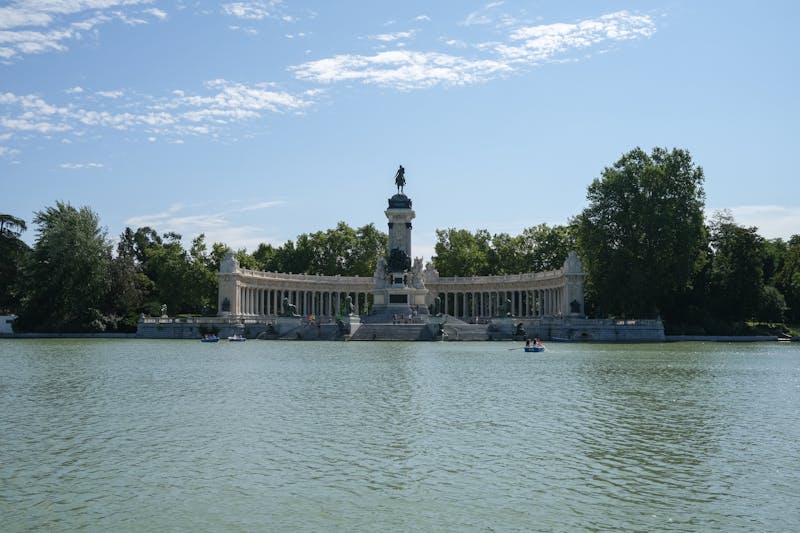 Scenic view of the monument in El Retiro Park, Madrid behind a tranquil pond on a sunny day.