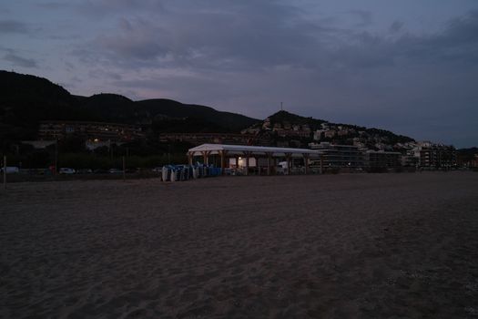 Serene beach scene at dusk with hillside buildings and a pavilion, exuding calm and tranquility.