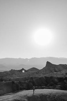 Black and white photo of a person exploring a vast, sunlit desert with mountains in the background.