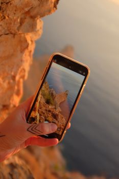Hand holding smartphone capturing scenic coastal cliff during sunset.