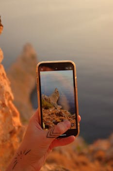 A hand holding a smartphone capturing a rocky seashore at sunset, showcasing natural beauty.
