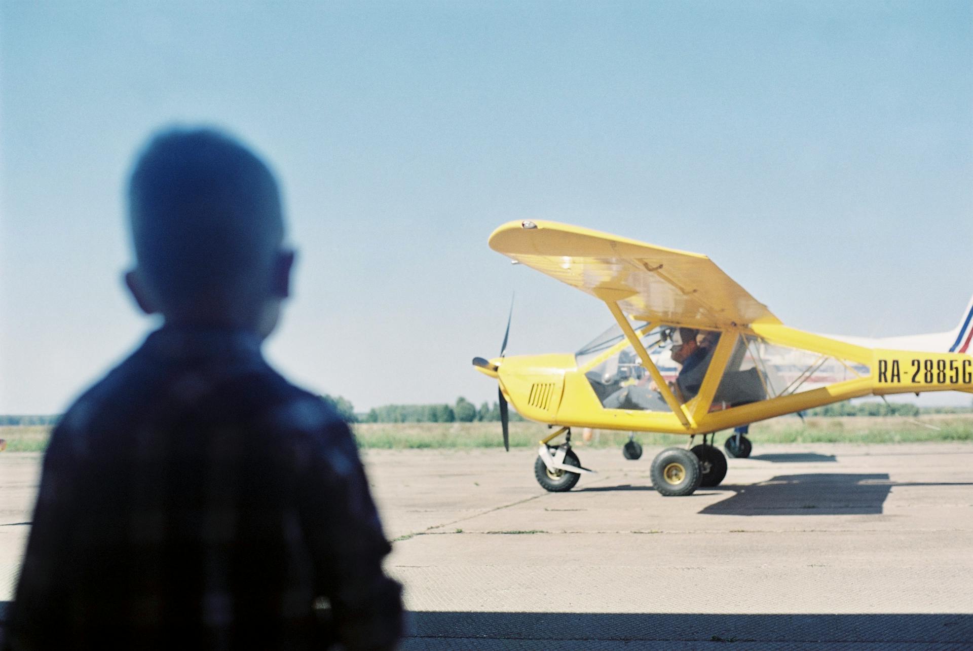 A young boy observes a small yellow aircraft on a clear runway, under a bright blue sky.