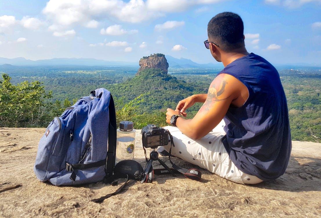 Free Man Sitting on Top of Gray Cliff Mountain Beside Backpack, Water Bottle, and Camera Stock Photo