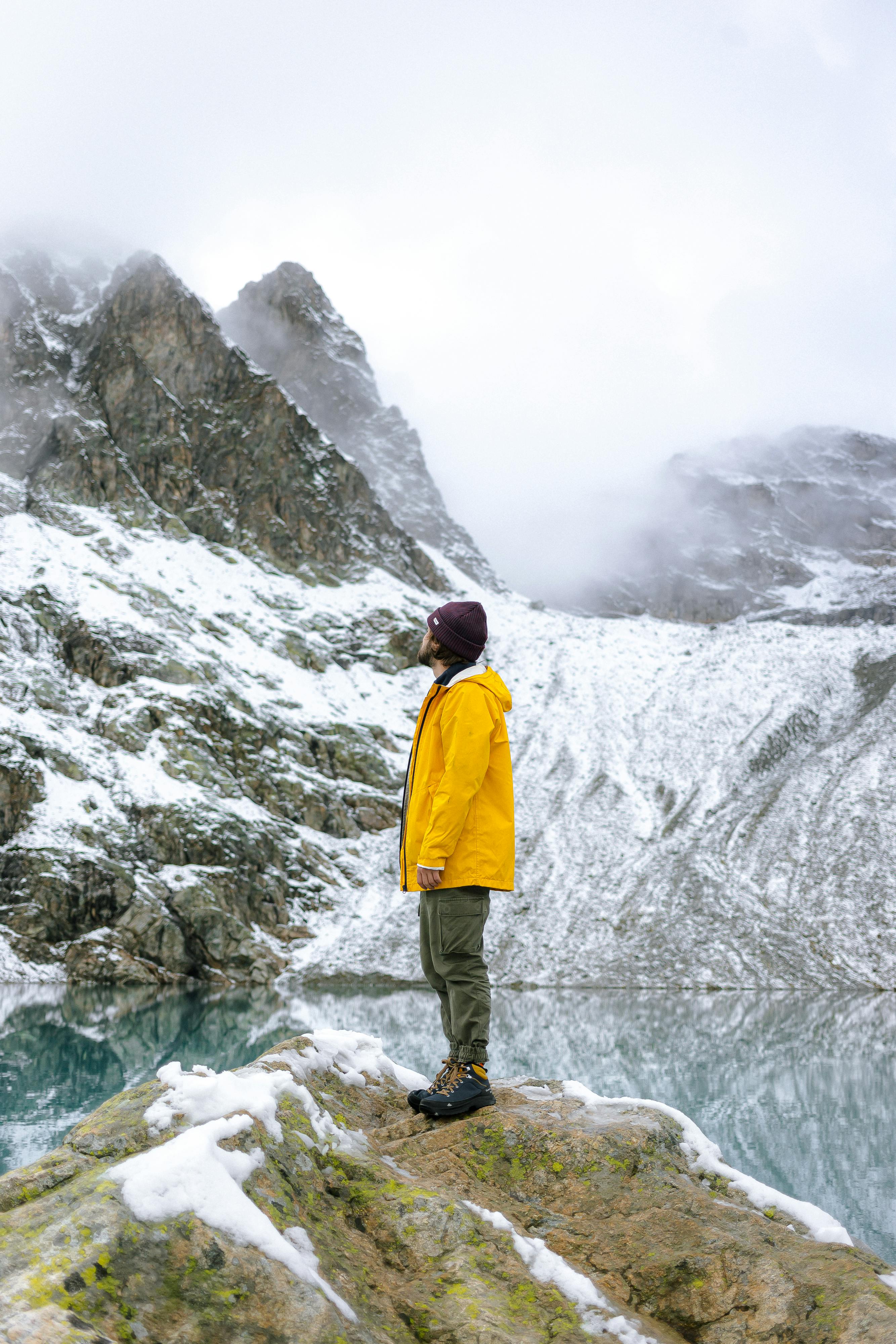 Man in yellow jacket standing in snowy mountain landscape, reflecting in a lake amidst misty weather.
