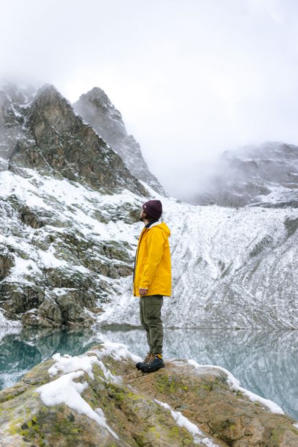 Man in yellow jacket standing in snowy mountain landscape, reflecting in a lake amidst misty weather.