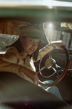 Woman in a vintage car interior, wearing a hat, looking at the camera.