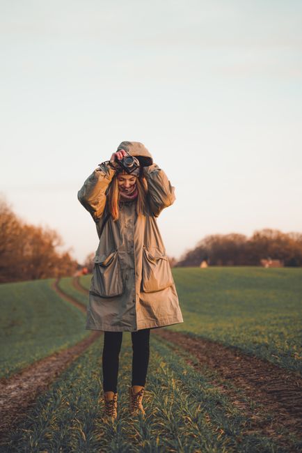 A woman wearing a raincoat stands in a grassy field during sunset, enjoying the peaceful outdoor atmosphere.