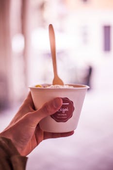 Close-up of a hand holding an ice cream cup with a wooden spoon, captured outdoors.