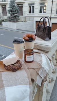 A stylish outdoor setup with coffee, pastries, candle, and handbag on a stone railing.