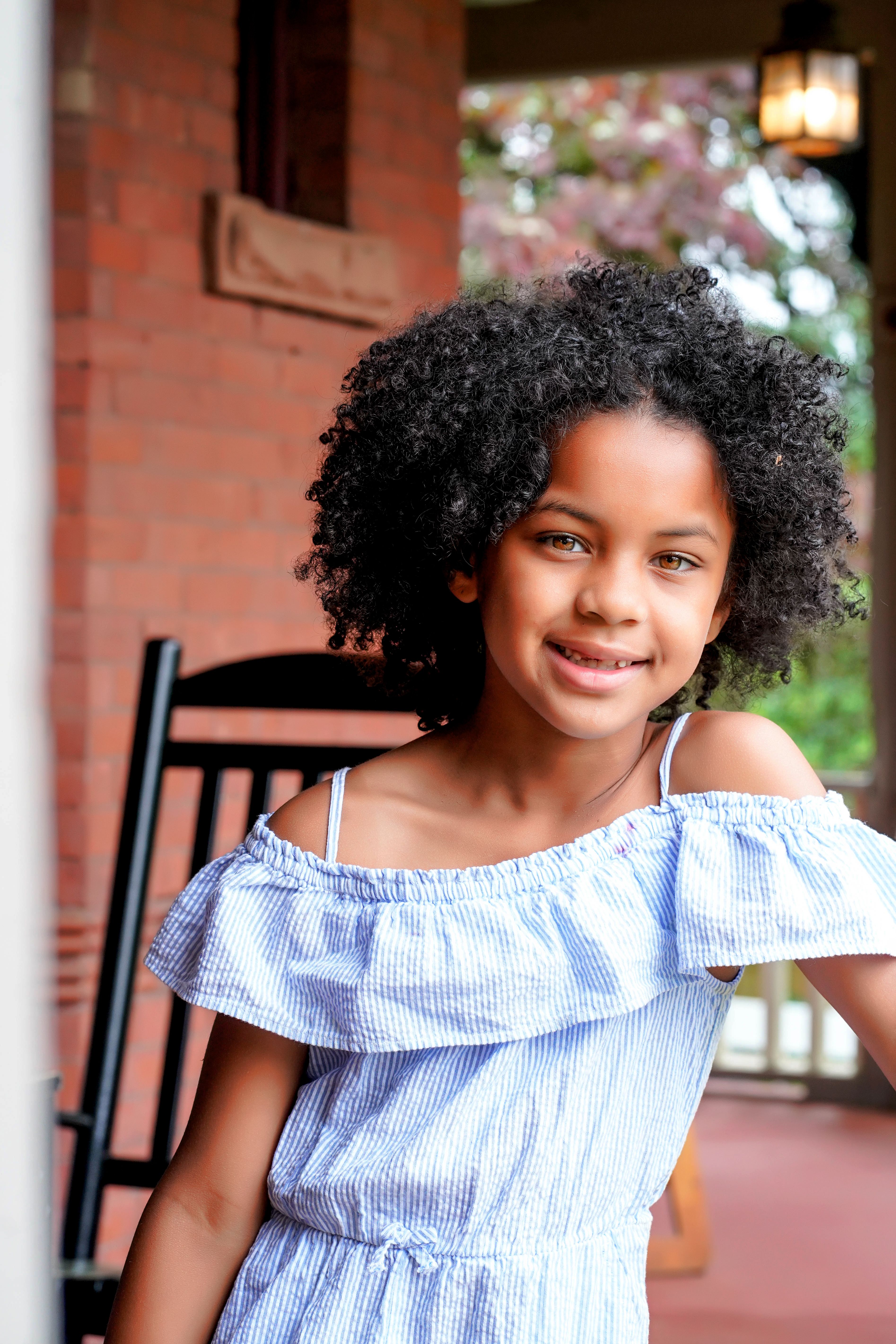 Adorable young girl with curly hair in a summer dress smiling on a porch.