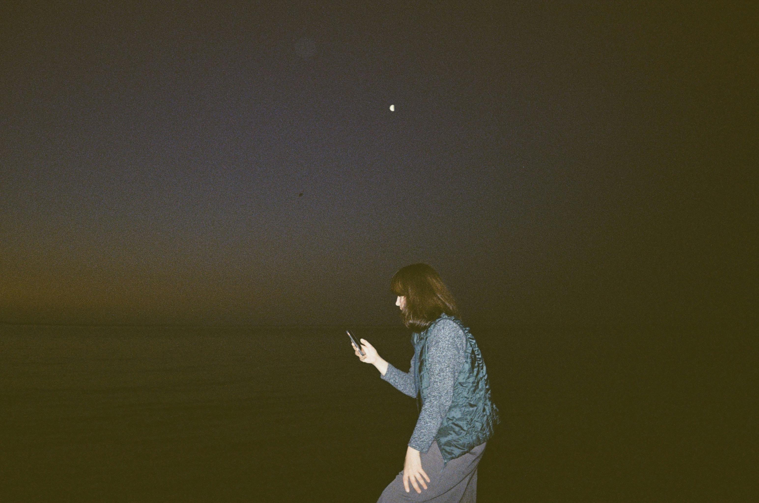 A woman with long brown hair using smartphone by the dark sea at night.