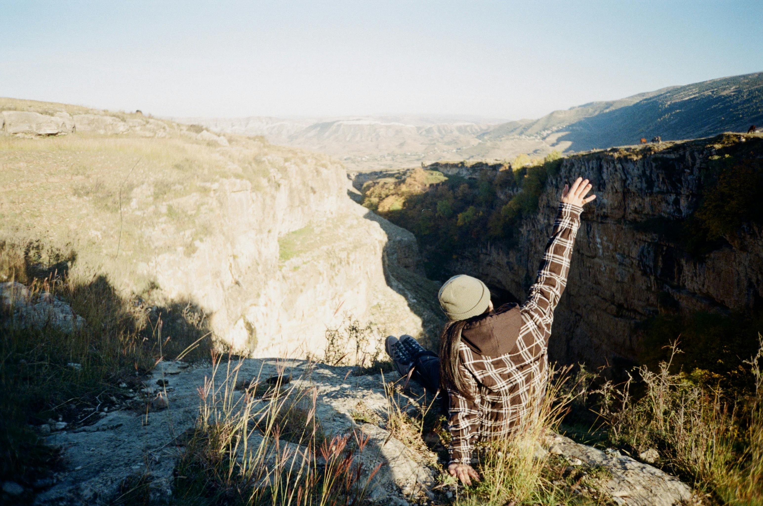 A woman sits on a cliff edge in a plaid shirt, raising her hand amidst a vast scenic landscape.