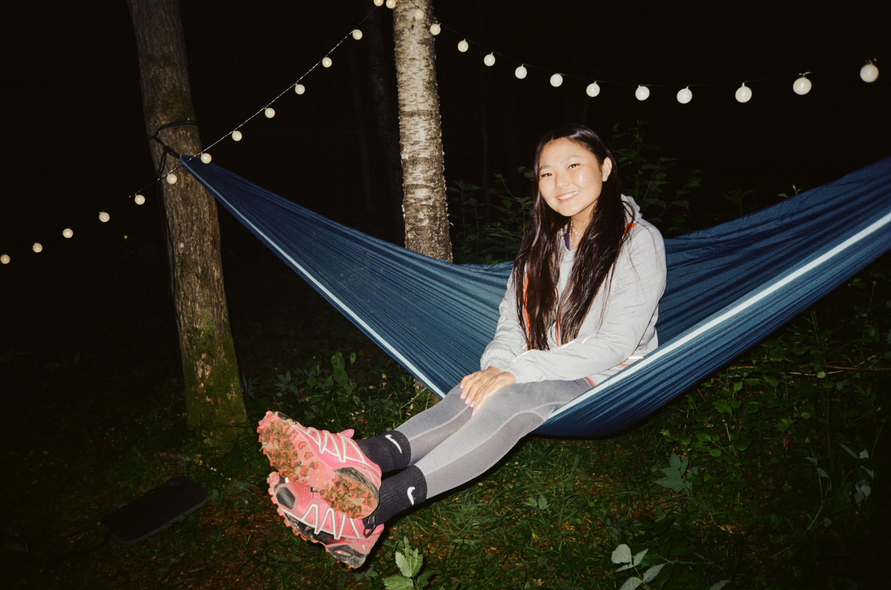 A young woman in sportswear relaxes in a hammock at night with decorative lights.