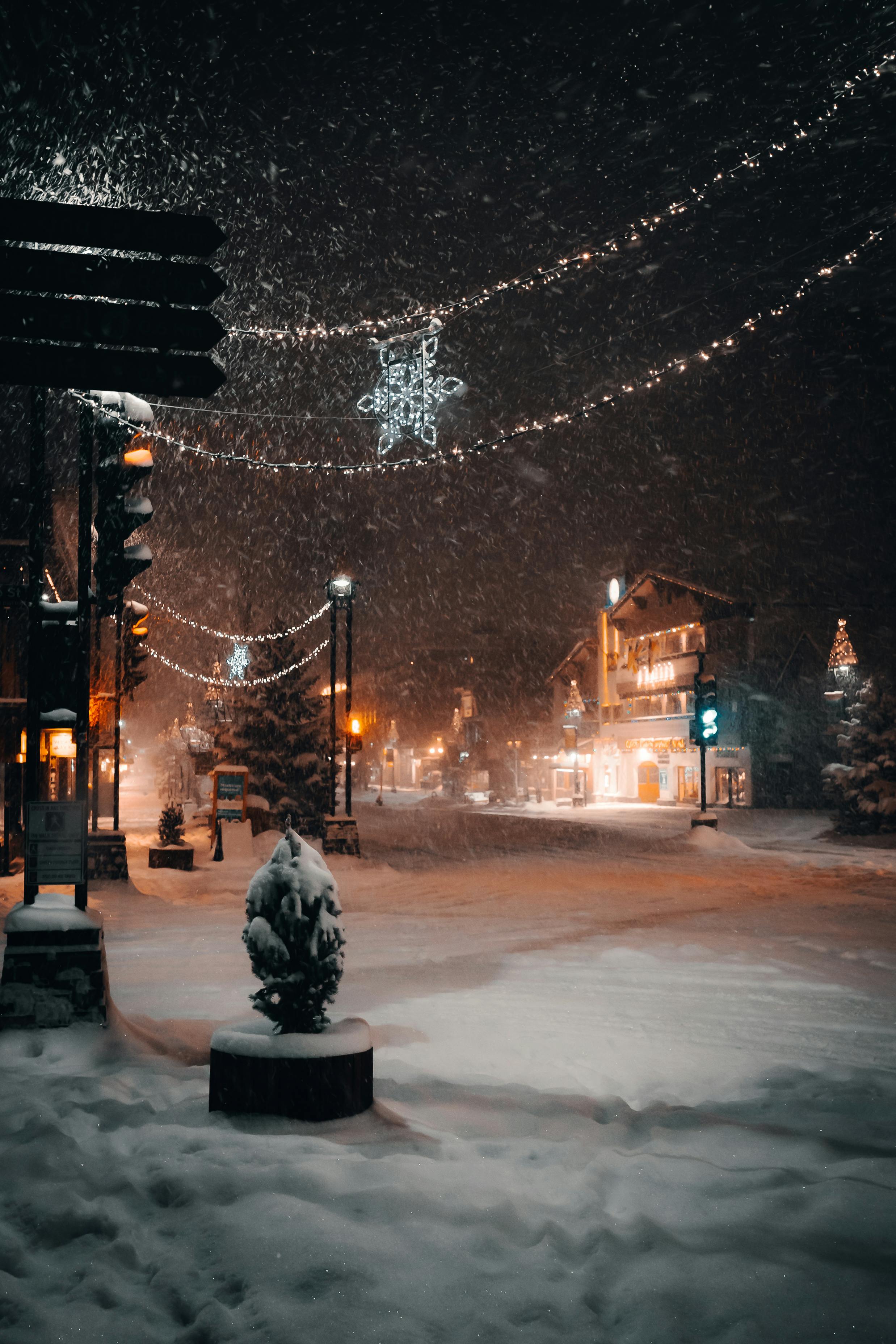 A picturesque snowy town square at night with Christmas lights and gentle snowfall creating a serene winter atmosphere.