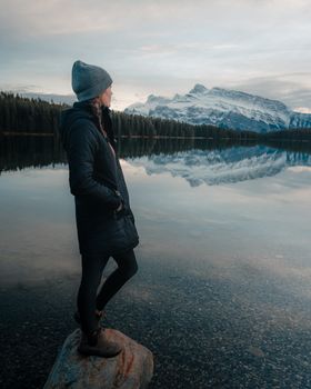 A solitary woman stands by a tranquil lake against a backdrop of snow-capped mountains in Alberta.