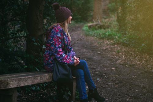 Free A young woman sits on a bench in a peaceful park, wearing warm, casual attire for a cool day. Stock Photo