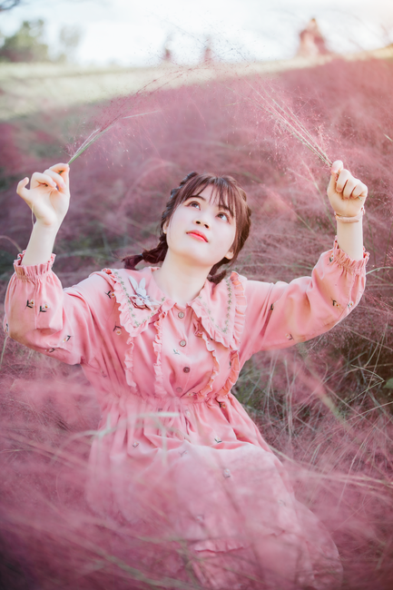 A young woman in a pink dress sitting amidst wispy pink grasses, looking up with a serene expression.