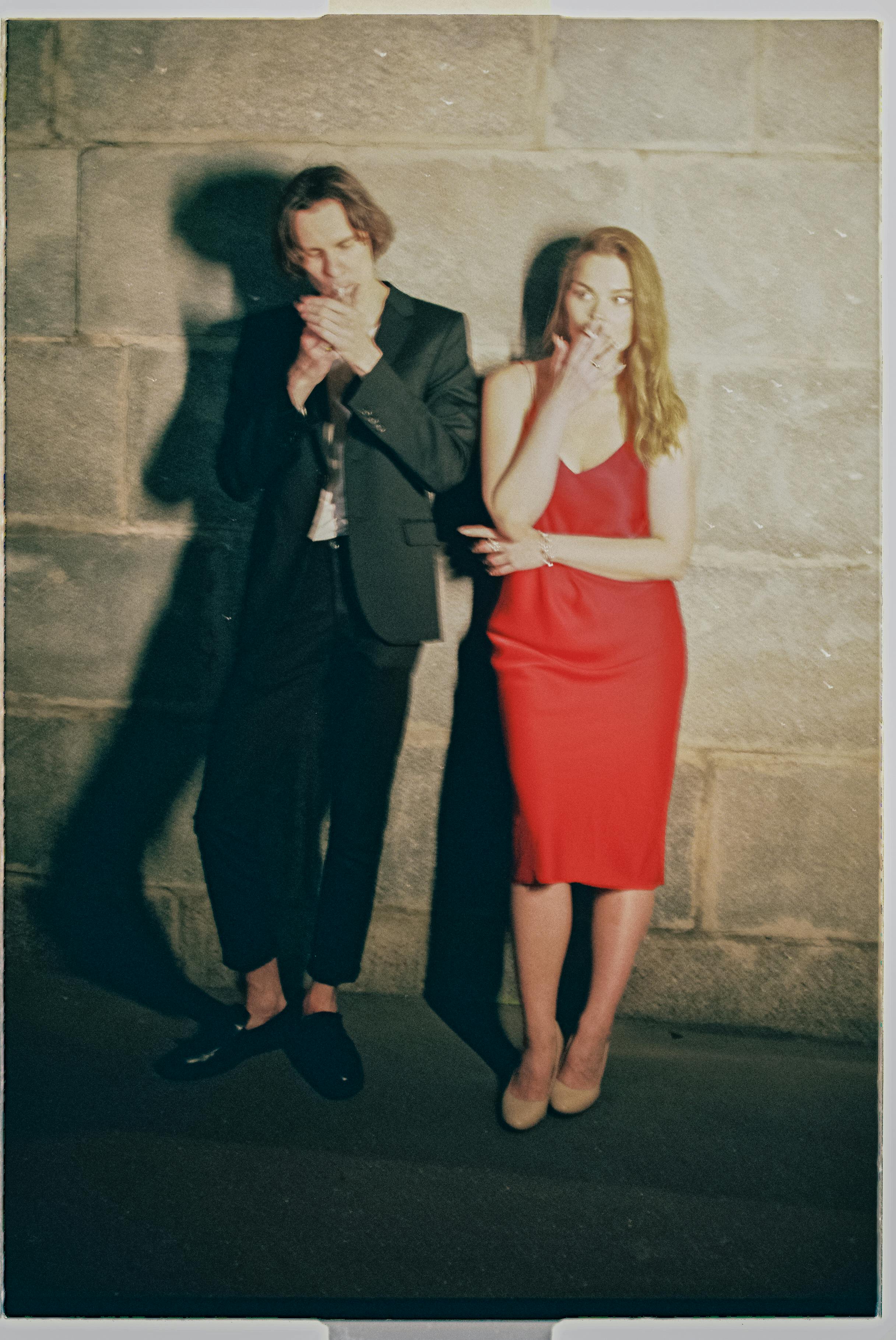 A stylish couple in formal attire smoking against a stone wall at night in Moscow, Russia.