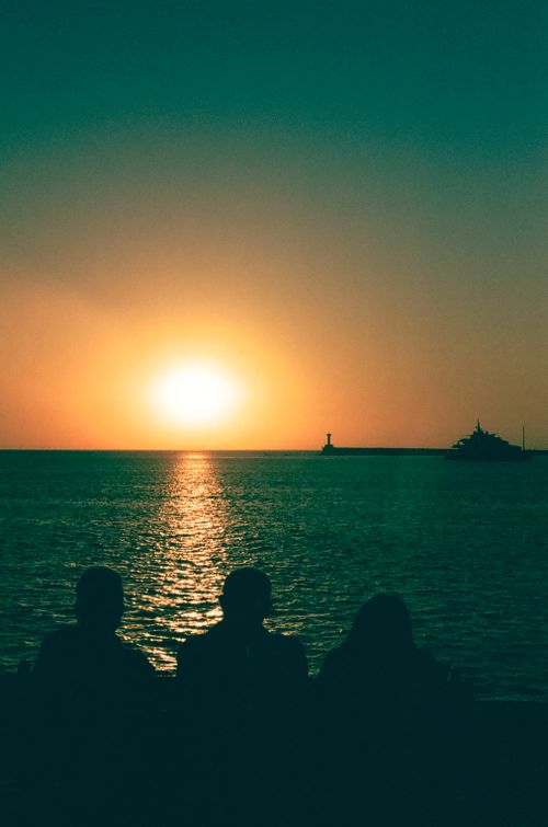 Free Silhouetted figures enjoy a breathtaking sunset over a calm sea with a distant lighthouse in view. Stock Photo