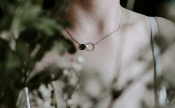 A close-up of a delicate silver necklace worn by a woman, surrounded by soft-focus foliage.
