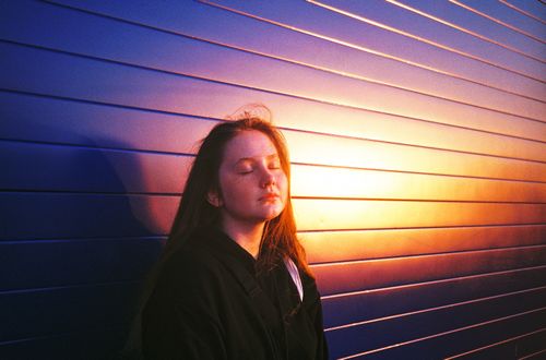 Free Teen girl with closed eyes enjoys a tranquil sunset against a vibrant wall. Stock Photo