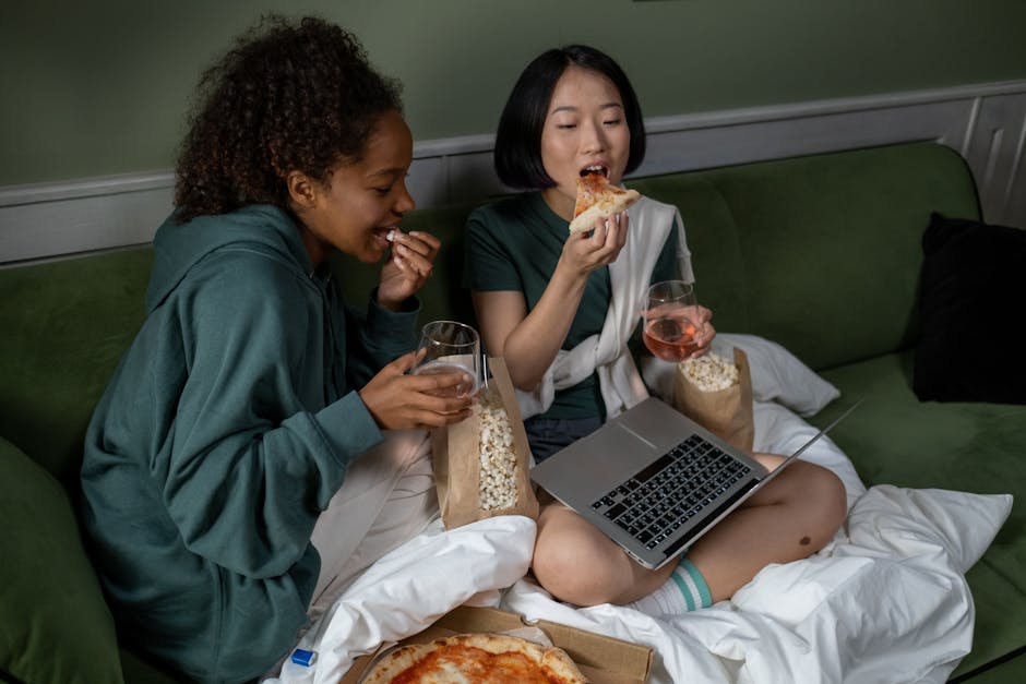 Two women relaxing indoors with pizza, popcorn, and drinks, watching a movie on a laptop.