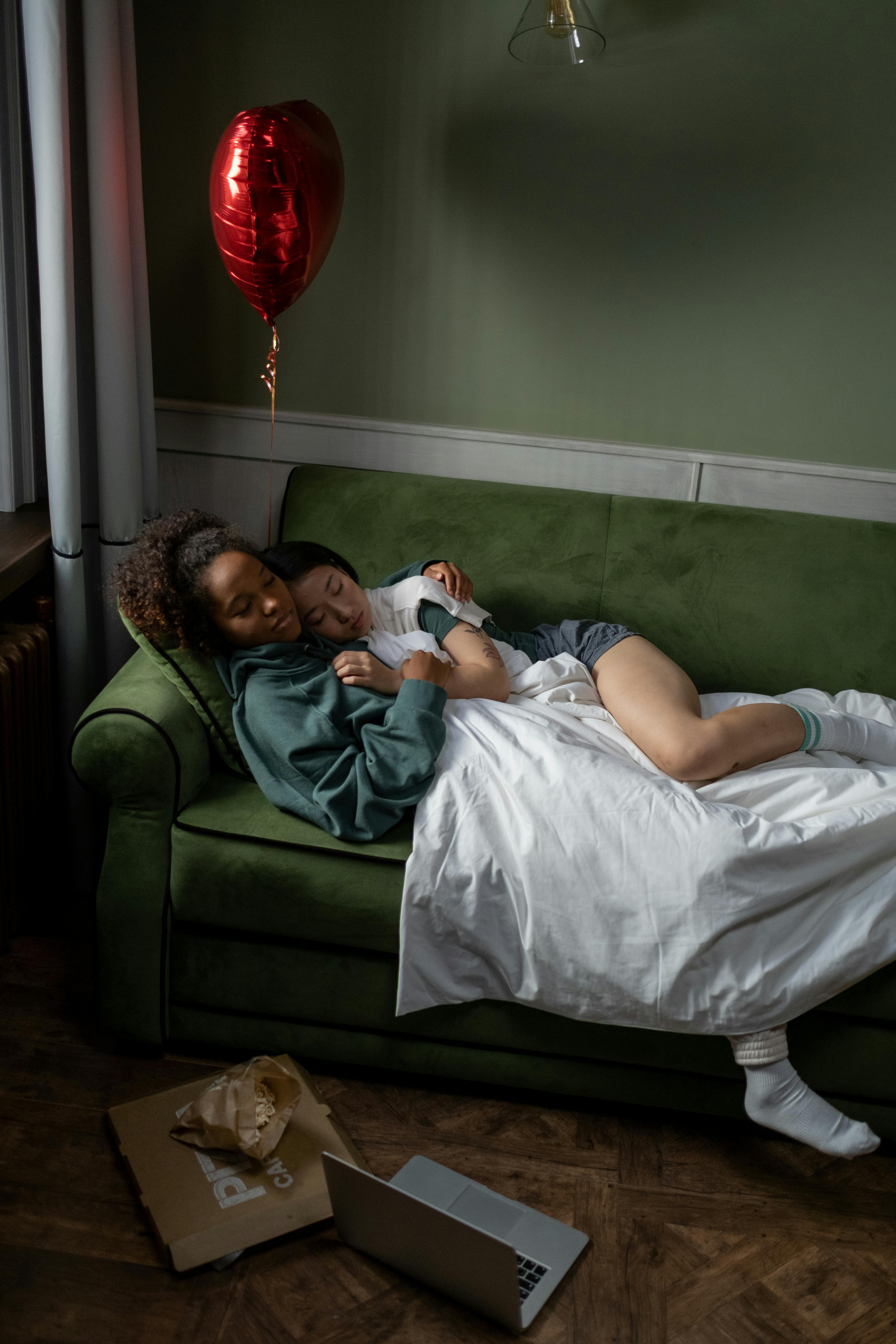 Couple cuddling on a green sofa with a heart balloon nearby, creating a cozy indoor scene.