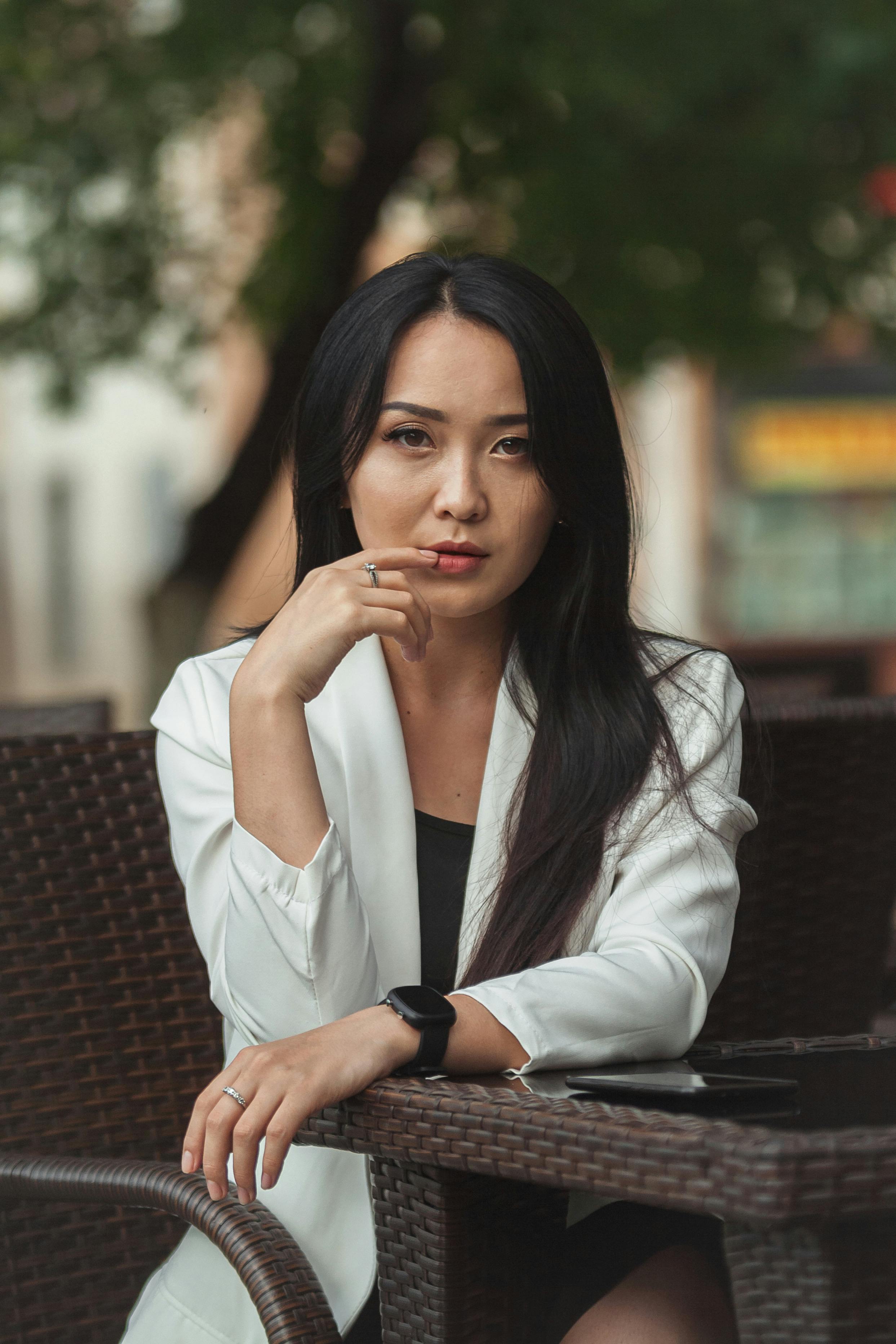 Elegant woman in a white blazer outdoors, focused in thought with a blurred background, exuding sophistication.