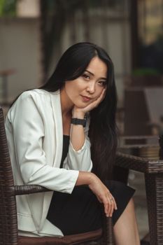 Portrait of a woman in a white blazer seated outdoors, exuding elegance and thoughtfulness.