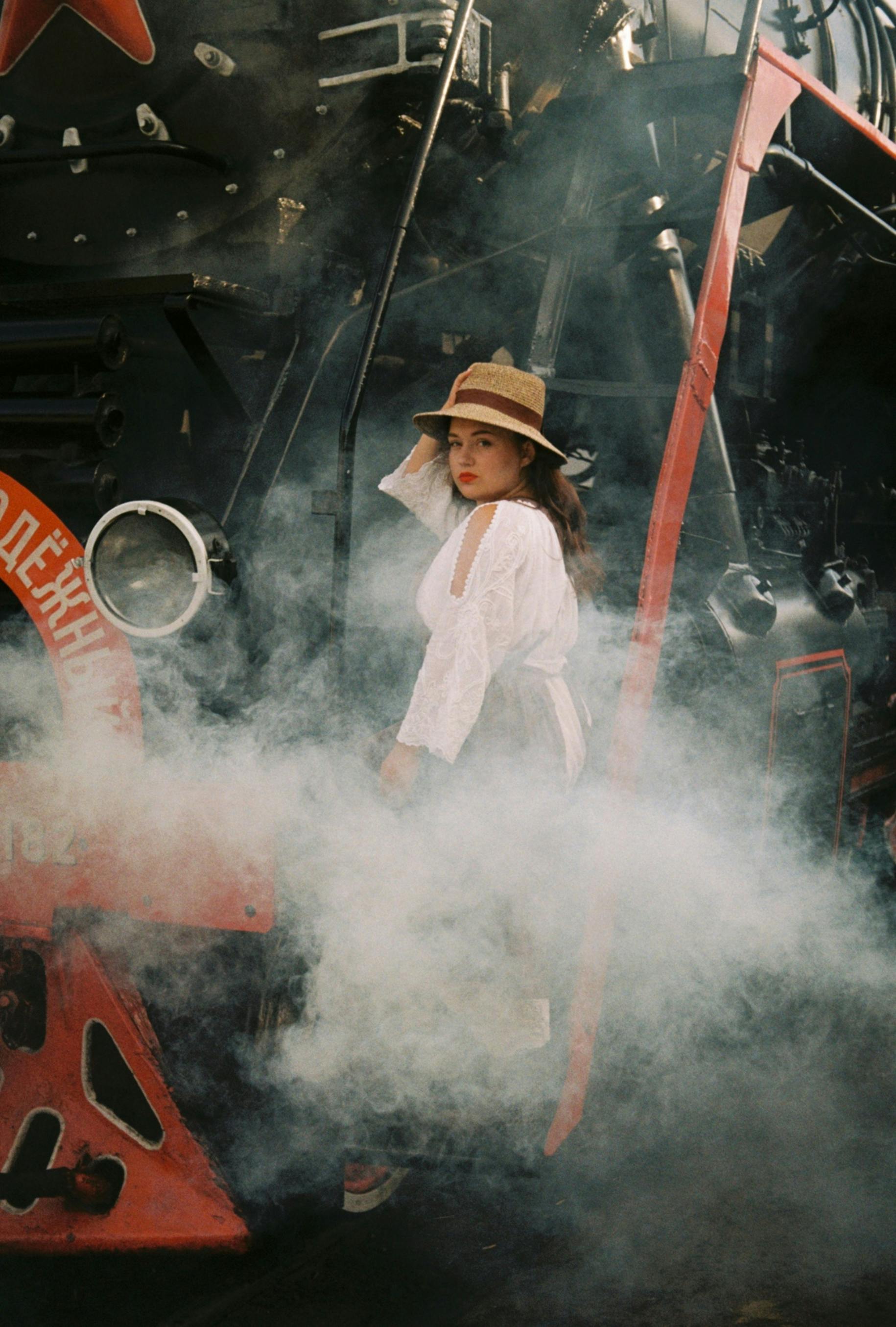 A woman in a white dress and hat standing near a smoky, vintage steam train.