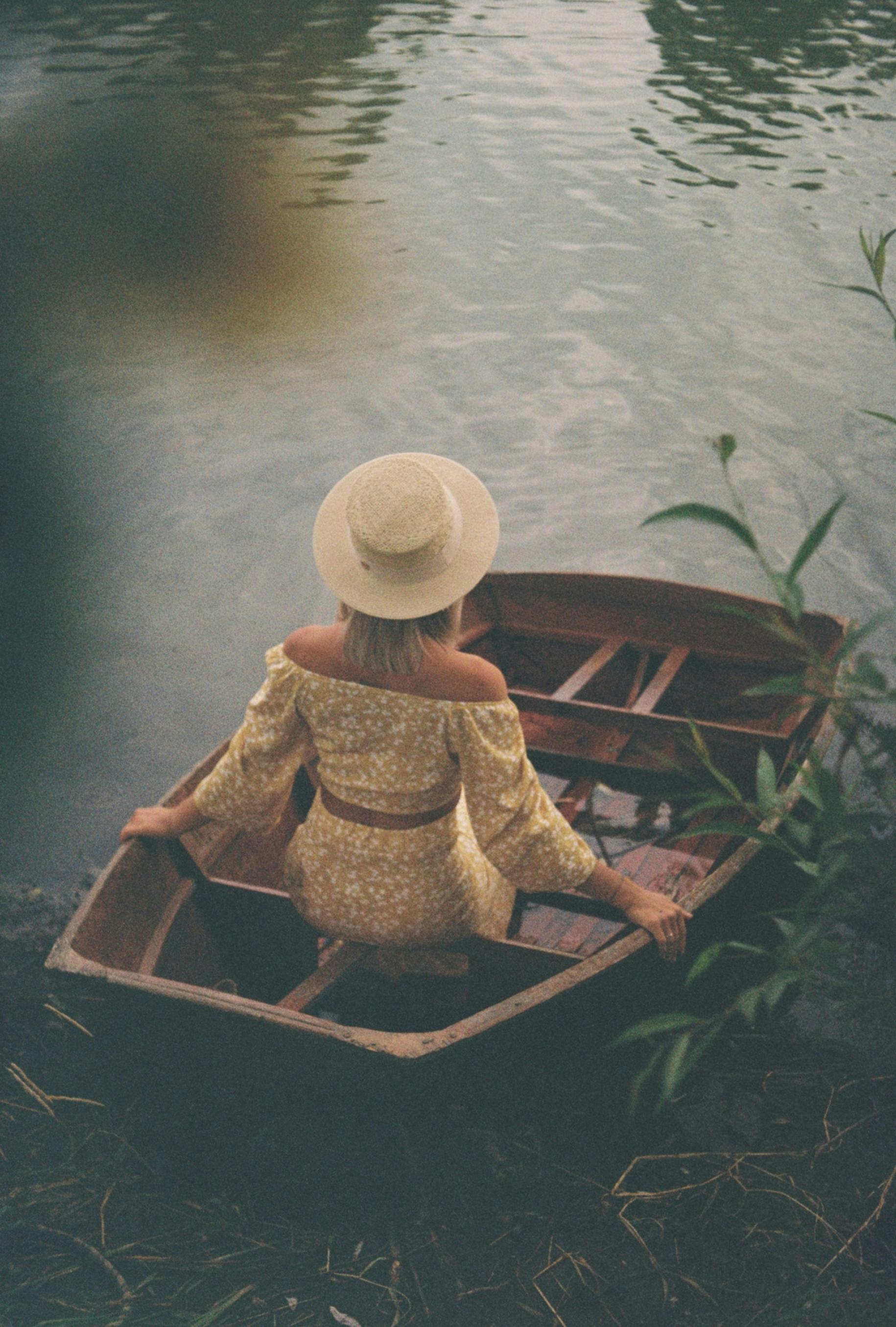 A woman in a hat sits in a wooden boat on a calm lake, evoking a peaceful outdoor scene.