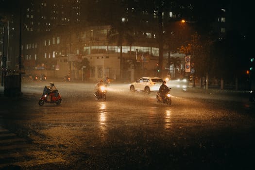 Motorcyclists and cars navigate through a rainy night on a city road, lights glistening.