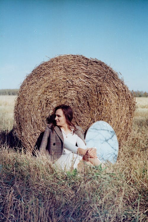 A Woman Wearing Brown Blazer Sitting on the Grass Field while Leaning on a Haystack