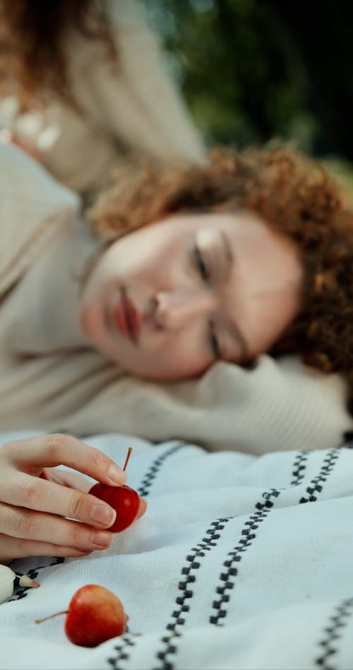 A Woman Holding a Crab Apple While Lying Down on a Picnic Blanket