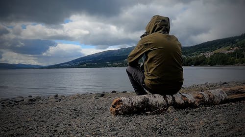 A Hooded Person Sitting on a Tree Log by a Lake