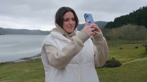 A Young Woman by a Lake Taking a Photo with her Smartphone 