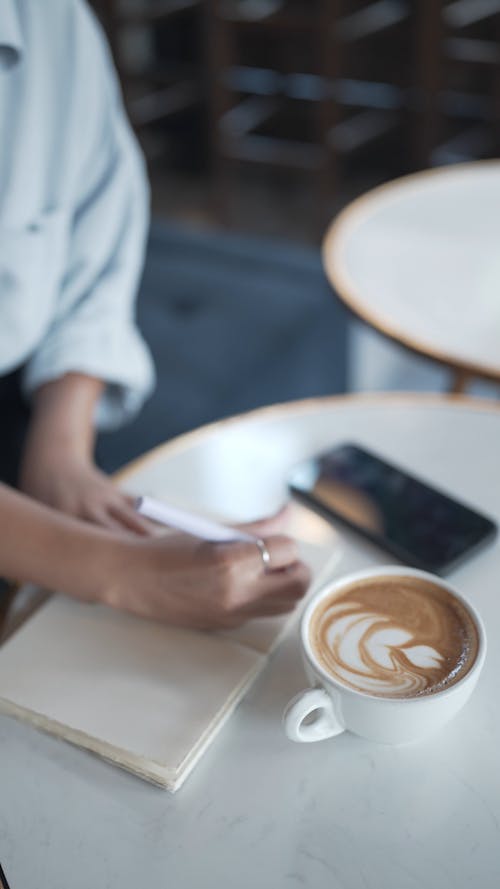 A Person Writing in a Notebook and Drinking a Cup of Coffee