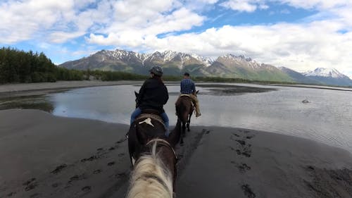 People Riding Horses through Lake near Mountains
