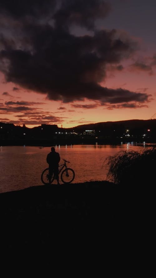 A Cyclist Standing by a Body of Water under a Twilight Sky